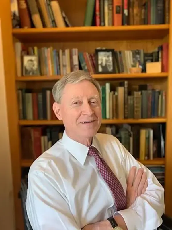 Professional portrait of a man with gray hair seated in front of a wooden bookshelf, wearing a light blue dress shirt and burgundy tie, with a calm expression.