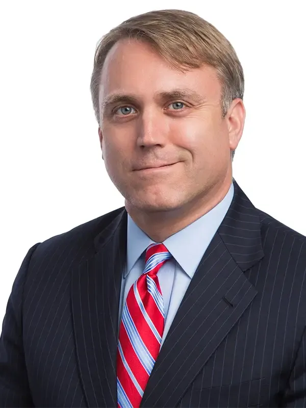 Professional headshot of a man with light brown hair, wearing a dark pinstriped suit, light blue dress shirt, and red and blue striped tie, smiling at the camera.