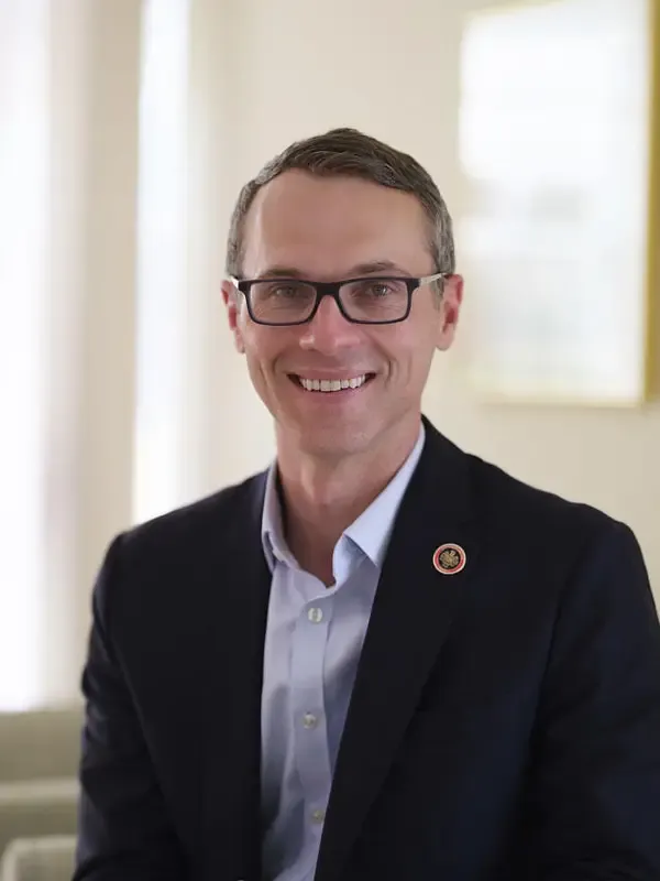 Man with dark hair and glasses wearing a navy blazer and light blue collared shirt, seated indoors against a neutral cream background, with a pin on his lapel.