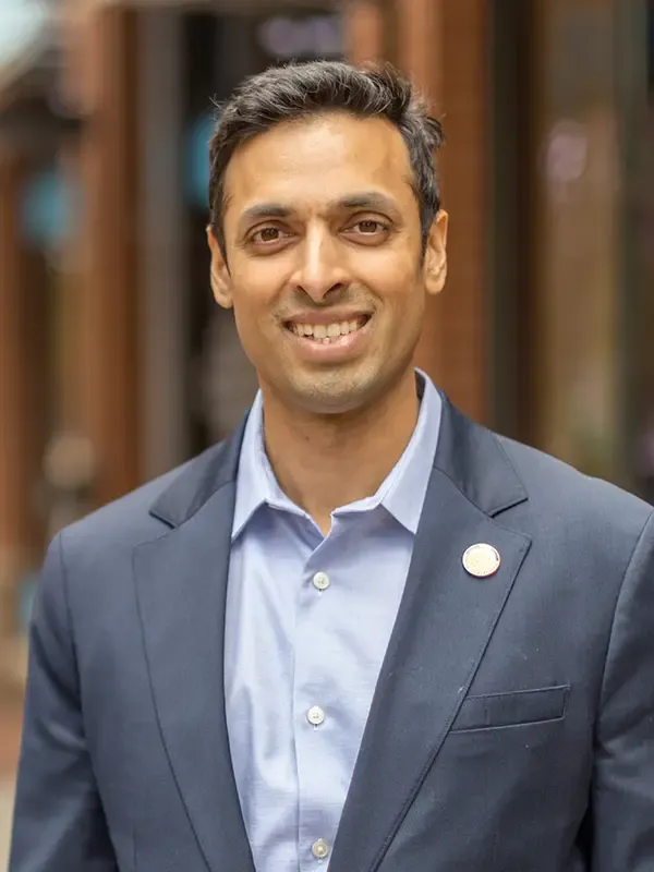 Man with dark hair wearing a gray blazer and light blue collared shirt, smiling warmly indoors against an urban architectural backdrop with warm brick tones.