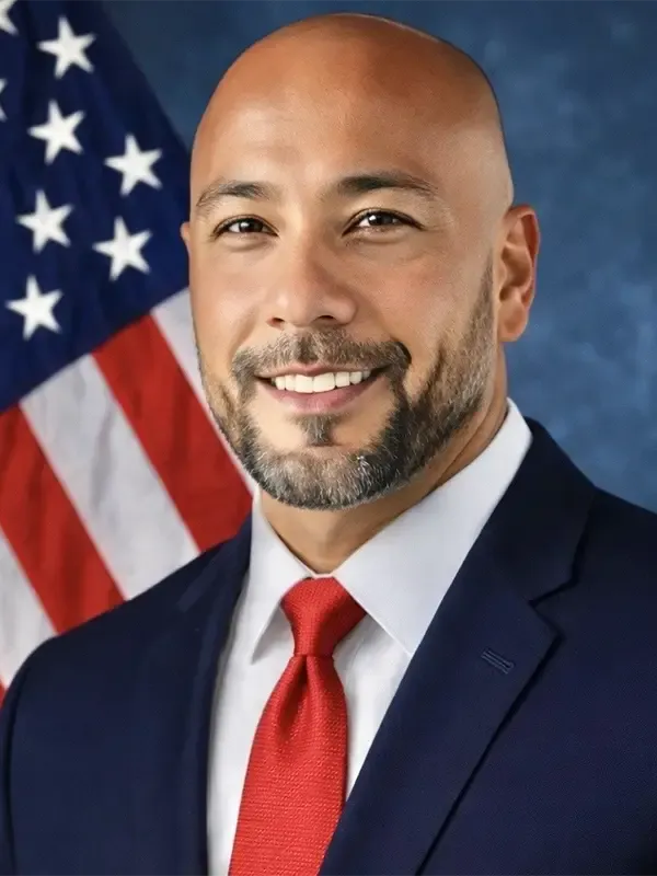 Professional headshot of a man smiling at the camera, wearing a dark navy suit, white dress shirt, and red tie, with a U.S. flag visible in the background.